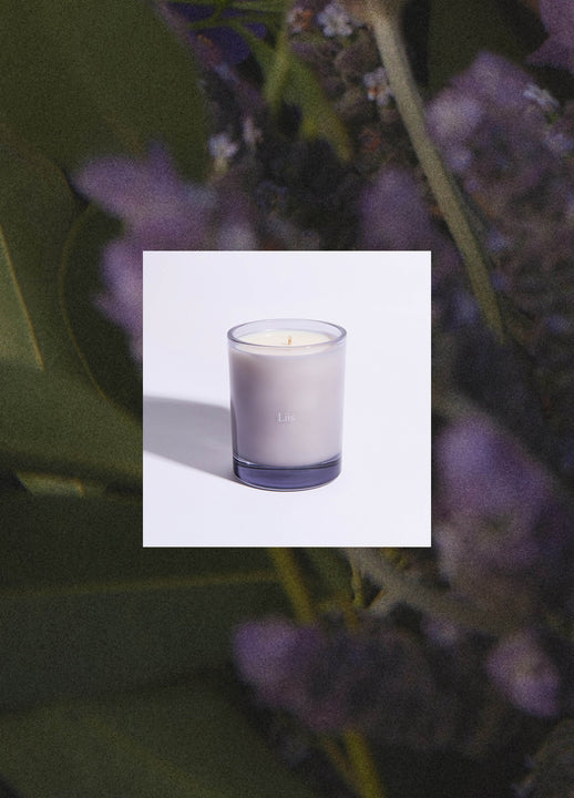 Load image into Gallery viewer, A single candle in a gray glass vessel centered on a white square overlay against a blurred background of green leaves and purple flowers.
