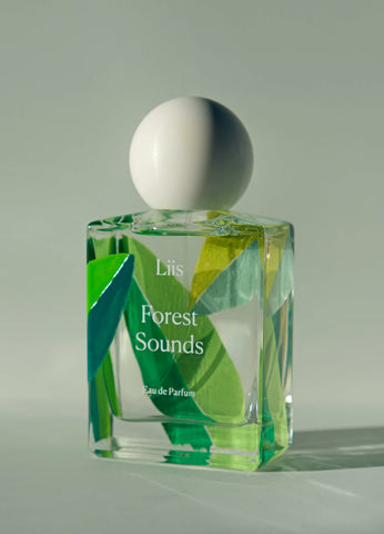 A clear glass perfume bottle with a white spherical cap, labeled 'Liis Forest Sounds Eau de Parfum', decorated with green and yellow leaf-like patterns, set against a neutral light gray background.